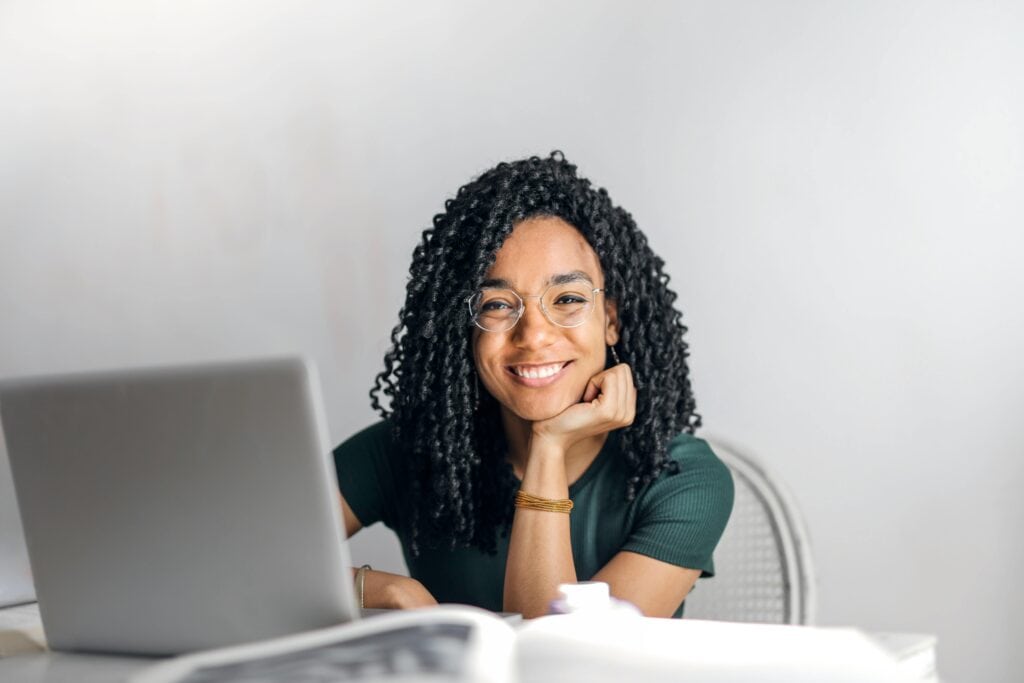Black woman with glasses resting her head on her hand smiling in front of laptop